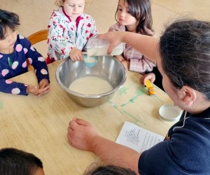 A group of children watching a teacher putting ingredients into a mixing bowl