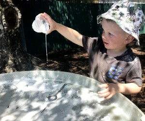 A young child playing with water, tipping water out of a container into a large tray